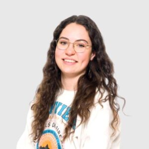 Jonge vrouw met krullend haar en een witte trui, glimlachend naar de camera, op een neutrale achtergrond.