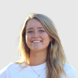 Jonge vrouw met lang, golvend haar en een glimlach, gekleed in een witte t-shirt, tegen een neutrale achtergrond.